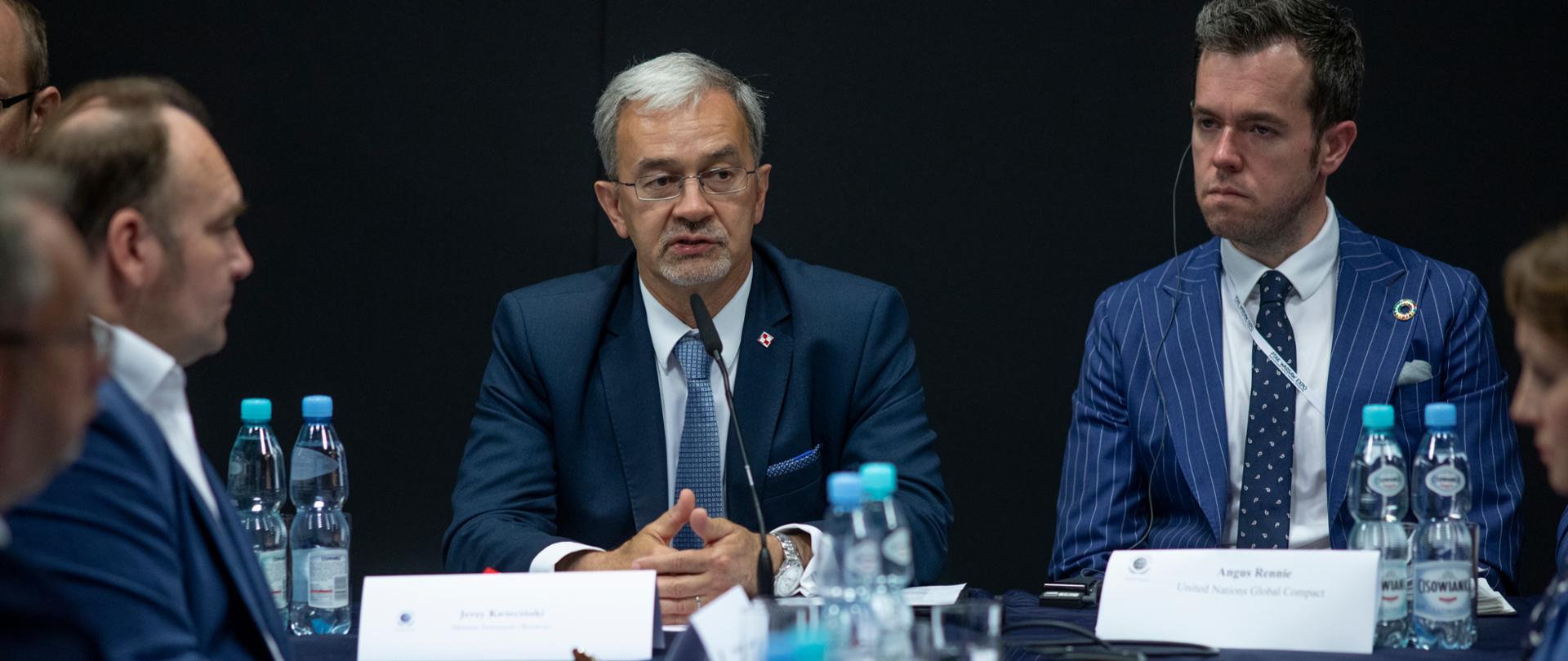 Grupa ludzi siedzi przy stołach. Na wprost minister Jerzy Kwieciński mówi do mikrofonu