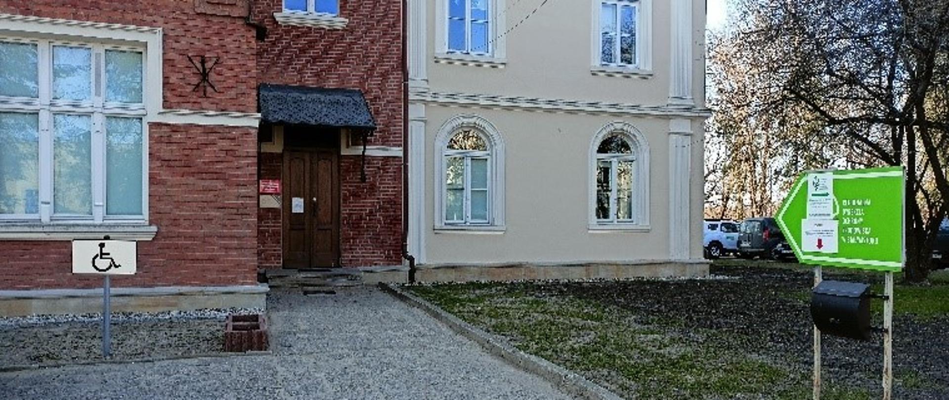 Na zdjęciu widnieje fragment budynku Regionalnej Dyrekcji Ochrony Środowiska z głównym wejściem do urzędu. Na pierwszym planie po lewej stronie zdjęcia widoczne jest miejsce parkingowe dla osób z niepełnosprawnościami. Po skrajnej prawej stronie widzimy tablicę informacyjną ze skrzynką podawczą. Tablica wskazuje drogę do urzędu