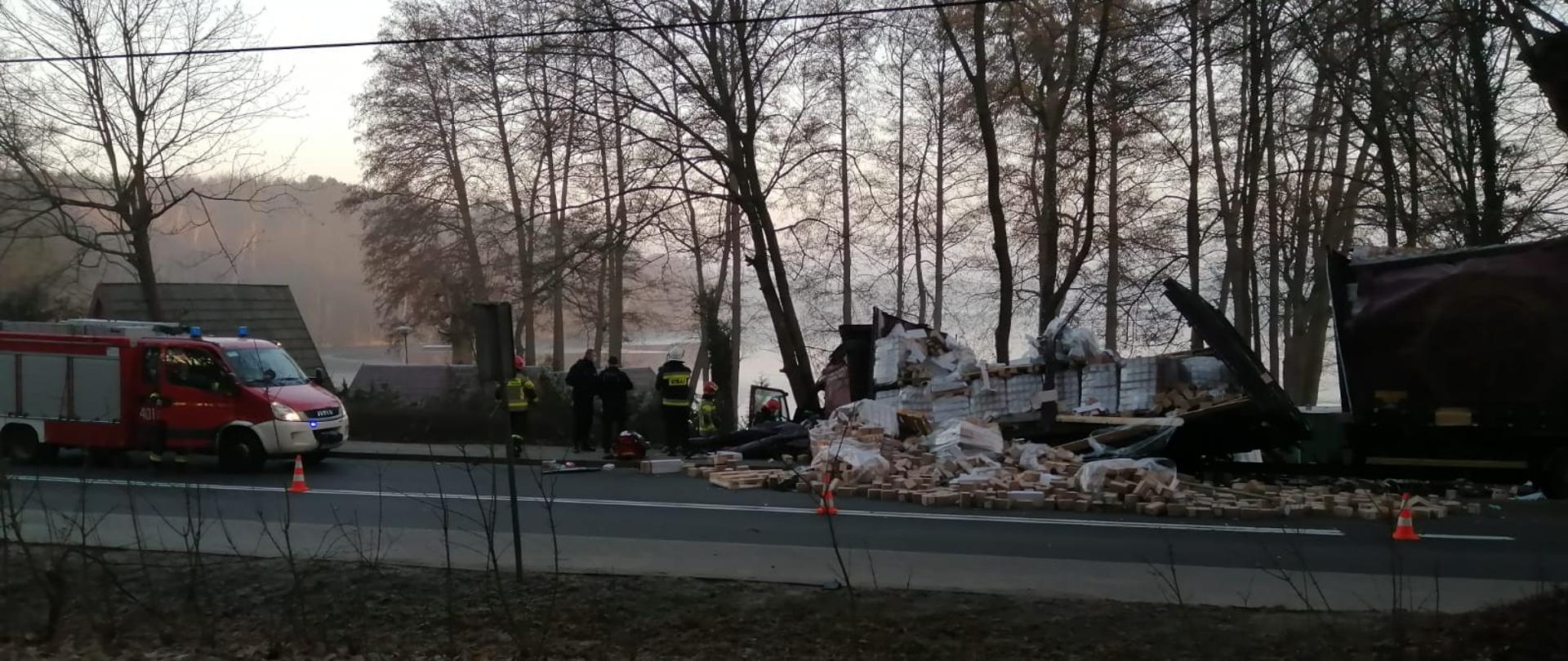 Zdjęcie przedstawia ujęcie odcinka drogi krajowej nr 22 w miejscowości długie, ujęcie wykonane prostopadle do przebiegu drogi. Po lewej stronie widoczny samochód ratownictwa technicznego z JRG PSP w Strzelcach Krajeńskich, w centralnej części samochód ciężarowy stojący na skarpie ze zniszczoną częścią ładunkową, zdartą plandeką, z częściowo rozsypanym ładunkiem z palet - ładunek stanowią niewielkie kartony , dalej po prawej stronie uszkodzona przyczepa samochodu ciężarowego. Na pasie jezdni w kierunku Strzelec Krajeńskich rozsypany ładunek z pierwszego członu zestawu. Między samochodem ciężarowym a pojazdem strażackim stoją tyłem strażacy w ubraniach specjalnych oraz dwóch funkcjonariuszy policji, dalej przy kabinie samochodu ciężarowego pracuje dwóch strażaków, nie widać co robią. W tle Drzewa porastające pobocze, dalej w dole widoczna tafla jeziora Lipie.