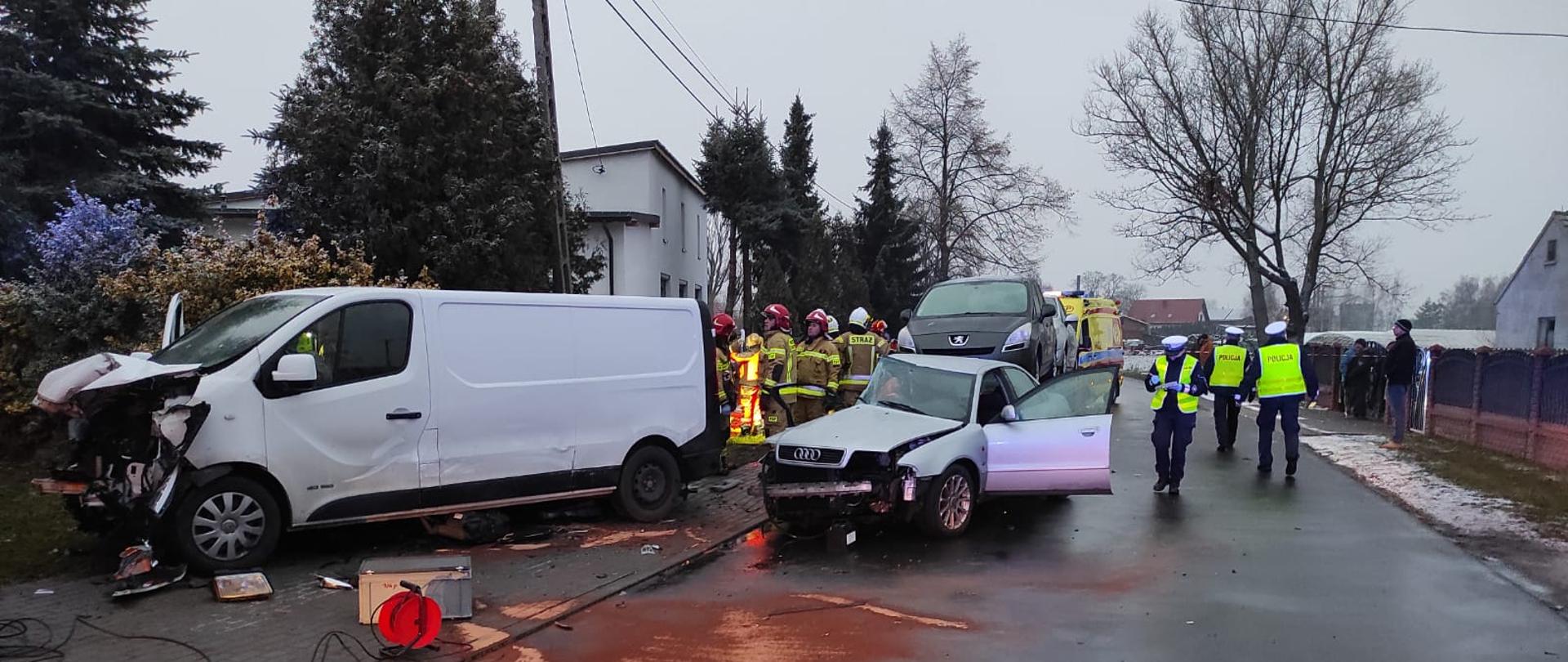 Na zdjęciu widać uszkodzone pojazdy, osobowy i dostawczy. Po prawej stronie od pojazdów budynek mieszkalny z betonowym płotem przed. Obok karetka pogotowia z sygnalizacją ostrzegawczą na dachu. W pobliżu funkcjonariusze Policji, PSP oraz osoby postronne. Prawa strona zdjęcia przedstawia betonowy płot w kolorze brązowym. W centralnym punkcie droga asfaltowa, posypana różowym sorbentem. Niebo zachmurzone. Pora zimowa.