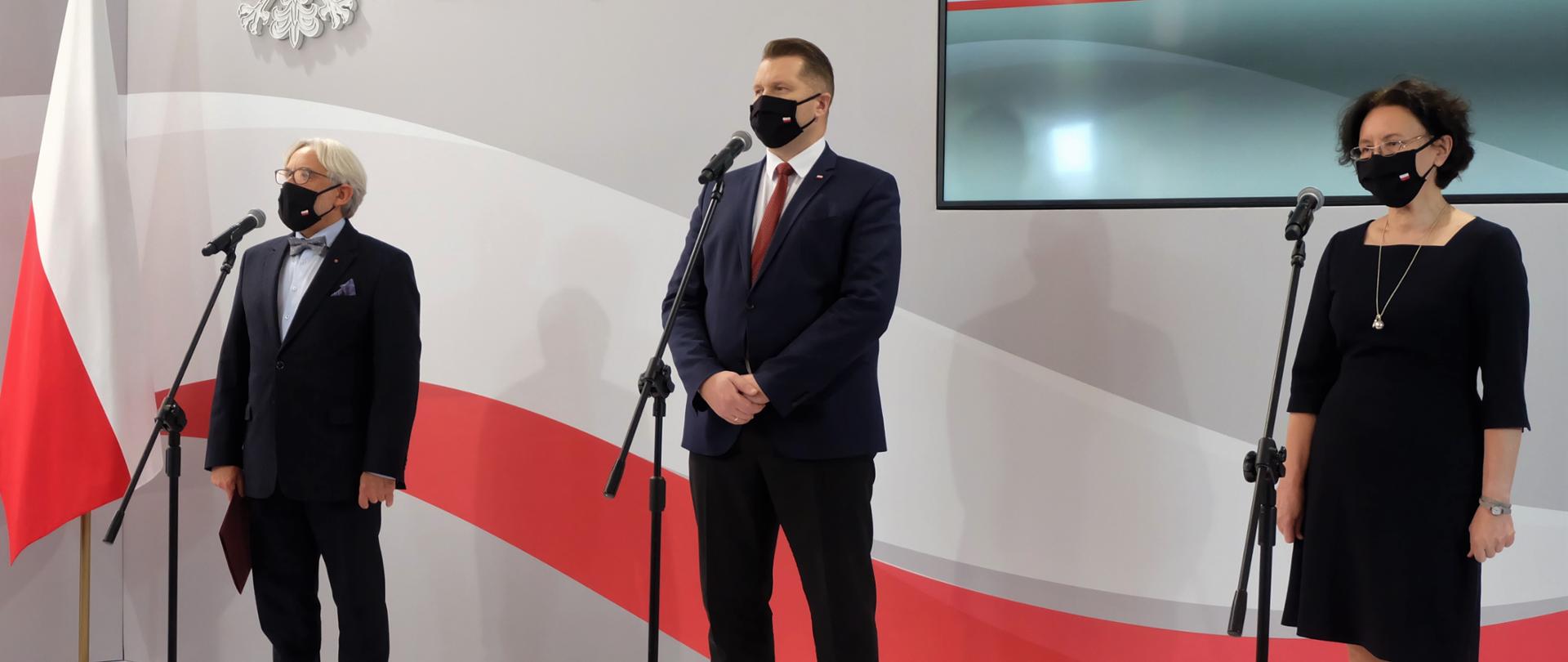 Na tle ściany z biało - czerwonym pasem stoją ministrowie i dyrektor NAWA, przed nimi mikrofony na stojakach.