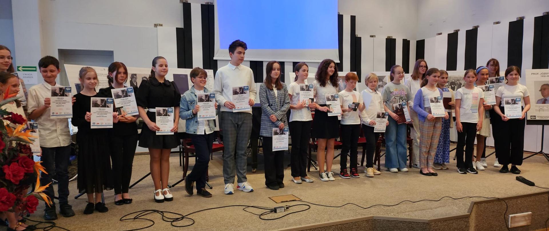 Dwadzieścioro jeden dzieci stoi na scenie Auli PSM w Jastrzębiu-Zdroju, wszyscy w rękach trzymają dyplomy.