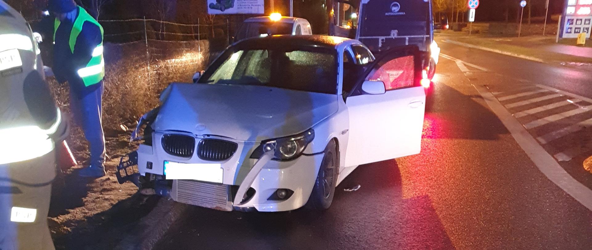 W centrum fotografii znajduje się uszkodzony samochód osobowy, który ma uchylone drzwi od kierowcy. Znajduje się on częściowo na jezdni i poboczu drogi. Uszkodzeniu uległa przednia lewa stron auta. Za pojazdem, w tylnej części fotografii znajduje się policyjny radiowóz. Przy samochodzie znajduje się technik policyjny oraz strażak. W oddali znajduje się stacja paliw. Zdjęcie wykonano w porze nocnej.