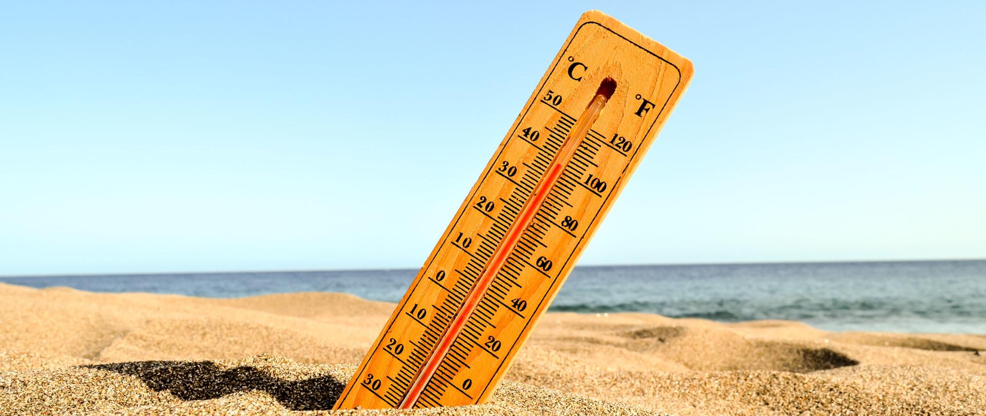 Zdjęcie przedstawia termometr wskazujący 40 stopni Celsjusza. Termometr jest wbity w piasek na plaży. W tle morze.