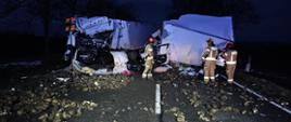 Na drodze wraki pojazdów doszczętnie zniszczonych w wyniku zderzenia czołowego na jezdni leżą rozsypane buraki cukrowe. Widać pracujących strażaków.