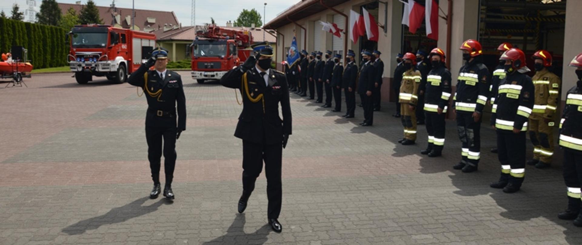 Zdjęcie przedstawia przegląd pododdziałów przez Świętokrzyskiego Komendanta Wojewódzkiego w asyście dowódcy uroczystości, pododdziały ustawione przed garażem JRG, w tle pojazdy pożarnicze.