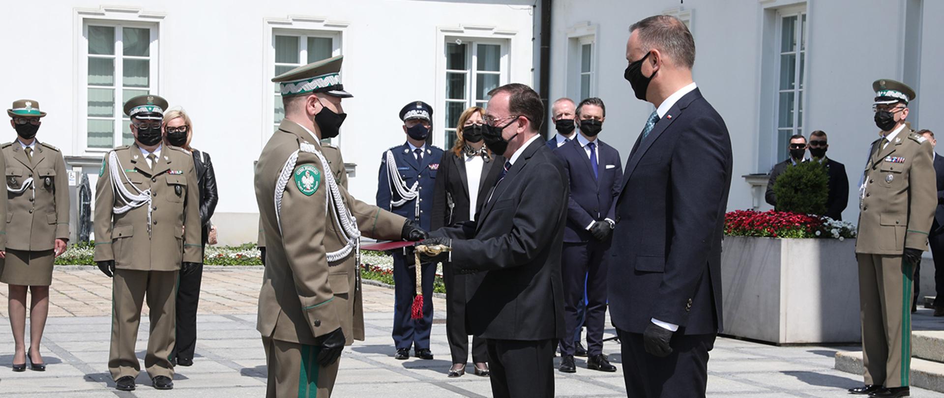 Minister Mariusz Kamiński na placu przed warszawskim Belwederem wręczający pamiątkową szablę nowo awansowanemu generałowi Straży Granicznej. Obok ministra Prezydent Rzeczypospolitej Polskiej Andrzej Duda.