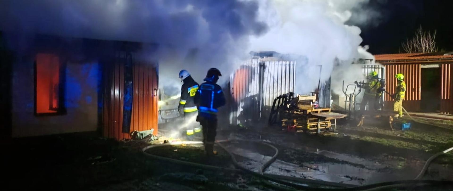 Zdjęcie przedstawia miejsce pożaru. W głębi budynek garażowy gdzie widać ogień i kłęby dymu, na tle garażu pracują strażacy.