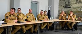 Za stołami ustawionym wzdłuż dużego pomieszczenia siedzą uczestnicy ćwiczeń, strażacy w mundurach piaskowych. Na ścianie z fototapeta z rodzajami broni palnej produkowanej na ZM