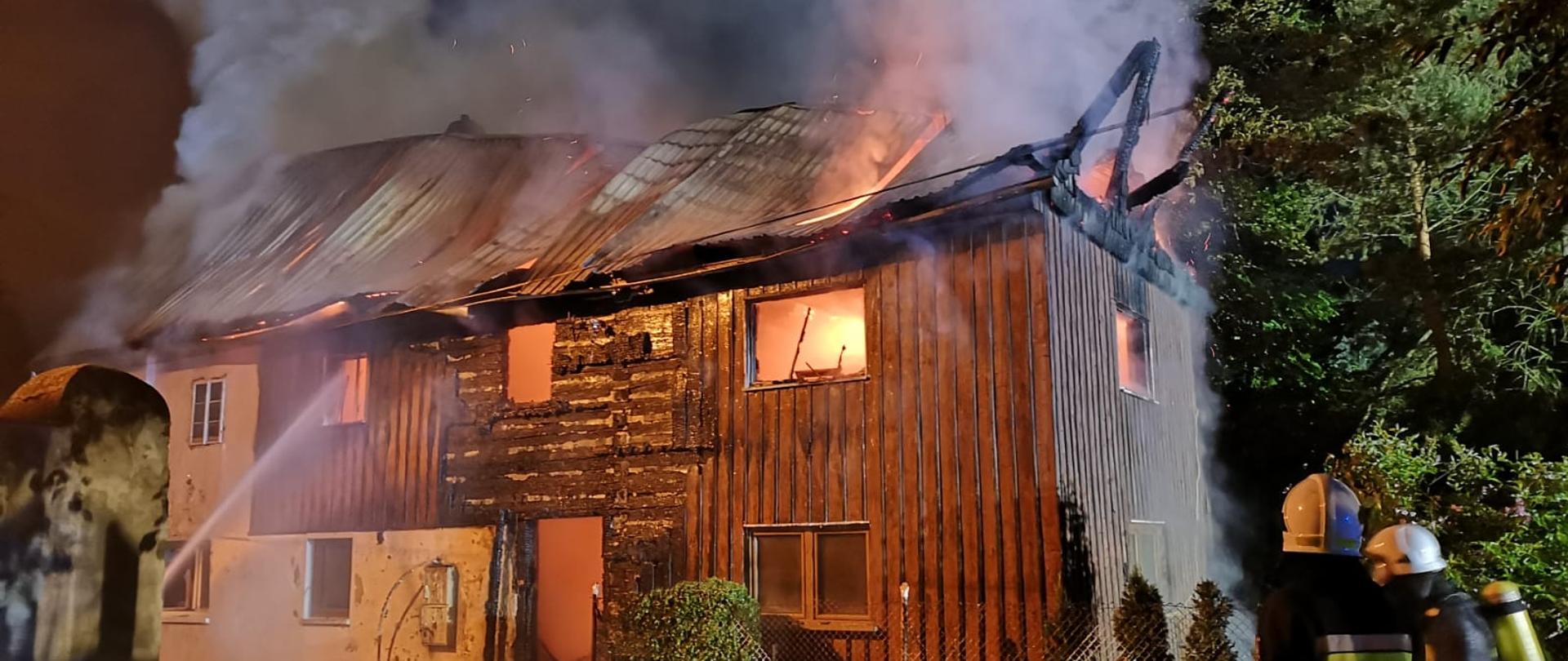 Pora nocna. Na głównym planie płonący drewniany budynek miieszkalny. W oknach na piętrze widoczne płomienie. Z dachu wydobywają się kłęby gęstego dymu.