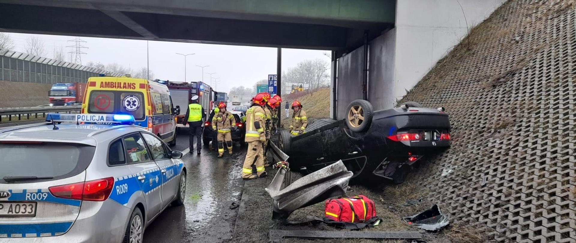 Zdjęcie przedstawia strażaków i policjantów interweniujących po wypadku samochodu osobowego na Drogowej Trasie Średnicowej w Rudzie Śląskiej. Na zdjęciu widoczny wrak pojazdu po dachowaniu, samochód pożarniczy, radiowóz policji oraz karetka pogotowia.
