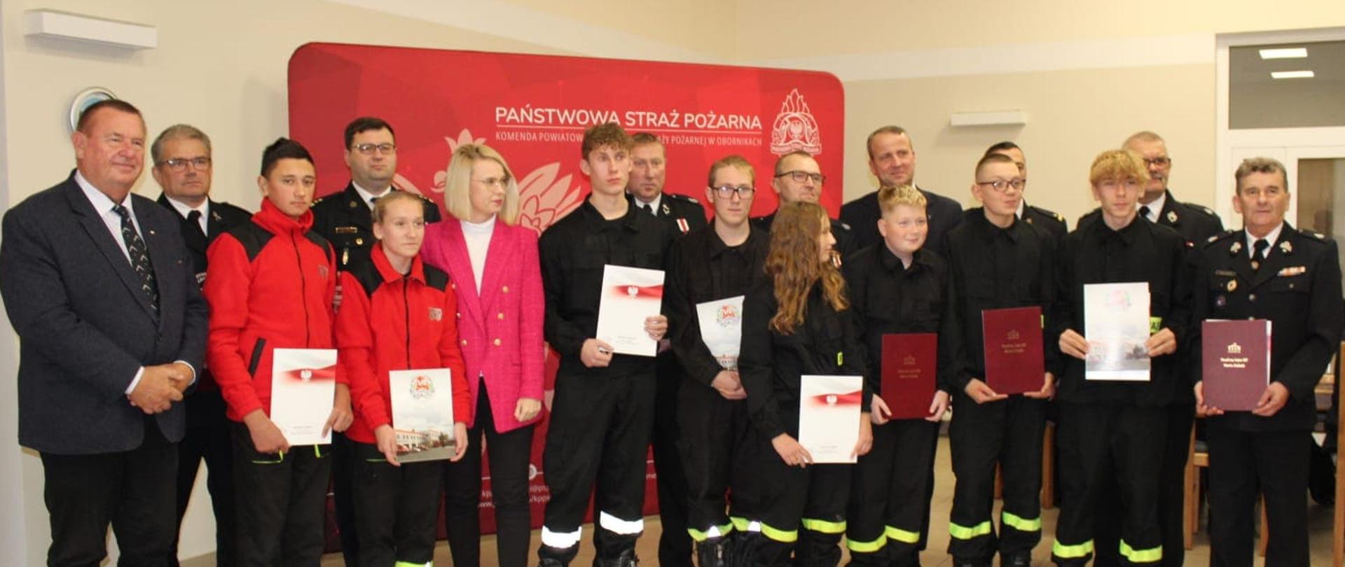 zdjecie grupowe małych strażaków i dorosłych na tle czerwonej ścianki