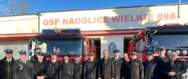 Grupa strażaków oraz osoby cywilne pozują do zdjęcia , w tle 2 samochody bojowe OSP oraz strażnica