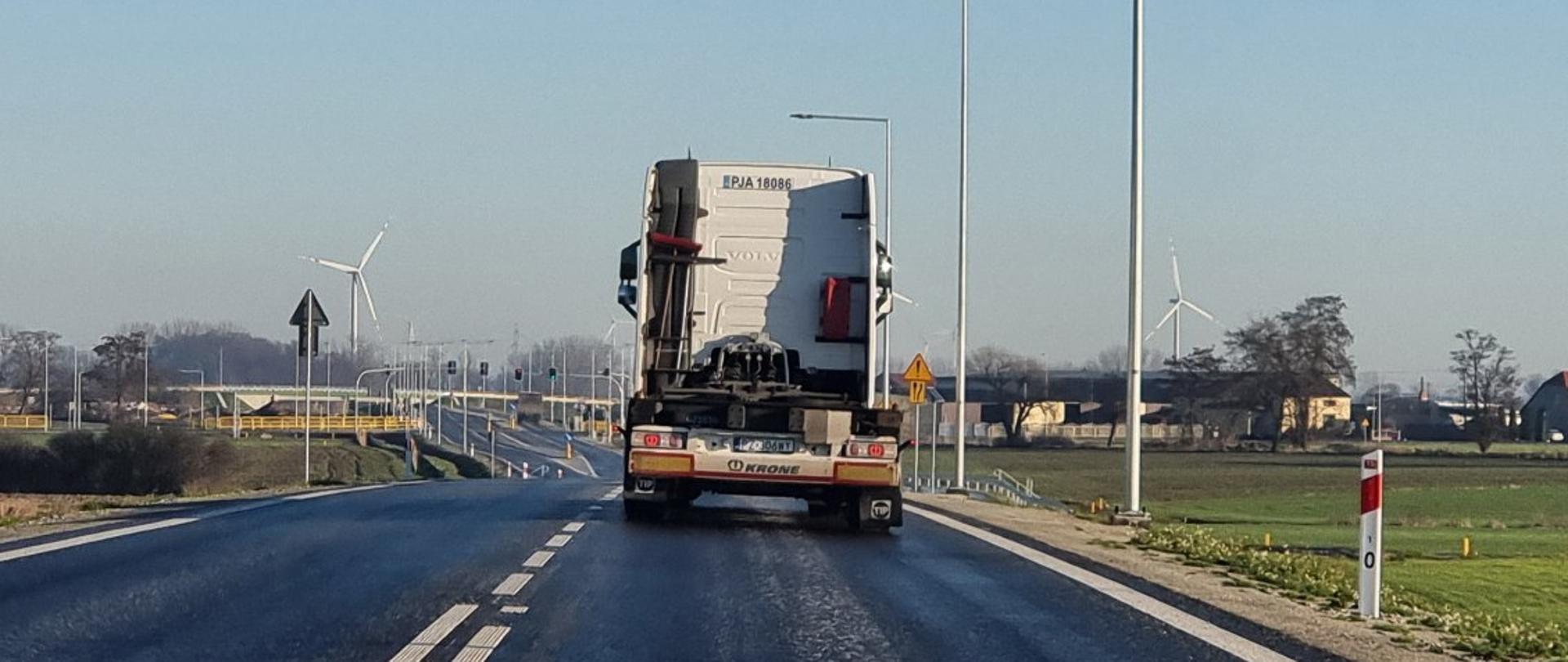 Widok nowej obwodnicy Koźmina – świeżo oddana do użytku droga z nowymi latarniami, biegnąca przez otwartą przestrzeń z widocznymi w tle turbinami wiatrowymi. Na jezdni porusza się ciężarówka przewożąca kabinę pojazdu, co symbolizuje rozpoczęcie ruchu na trasie.