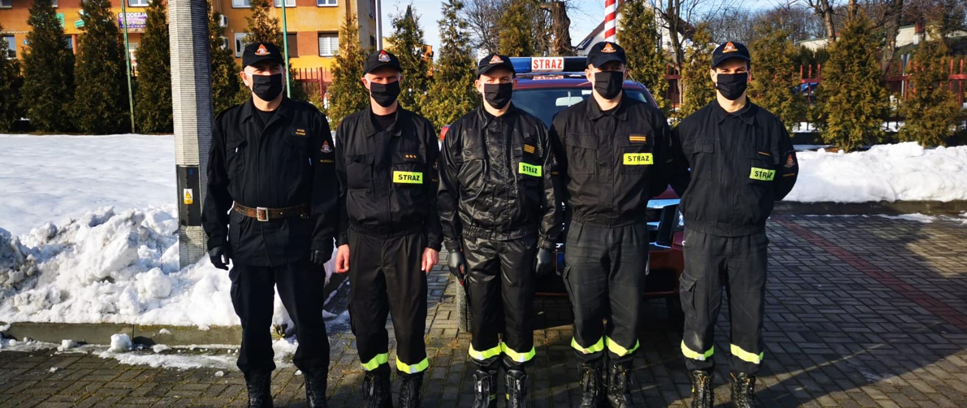 Zdjęcie przedstawia 5 strażaków ubranych w czarne ubrania koszarowe stojących na kostce brukowej przed samochodem służbowym jest to grupa z KP PSP Włoszczowy