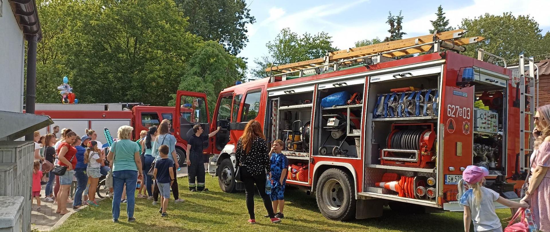 Zdjęcie przedstawia dwa wozy strażackie. Pomiędzy autami dorośli i dzieci przyglądają się wozom strażackim.