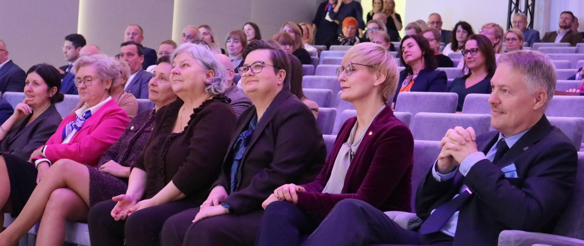 Kilka osób siedzi w pierwszym rzędzie na szarych fotelach w audytorium, obserwując wydarzenie. Widoczne są różnice w ubiorze, od garniturów po bardziej casualowe stroje.