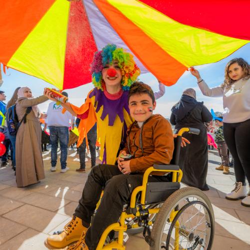 Uśmiechnięty chłopiec na wózku inwalidzkim z namalowaną flagą Palestyny na policzku. Obok niech uśmiechnięty kolorowy klałn. Wokół nich śmiejący się ludzie. 