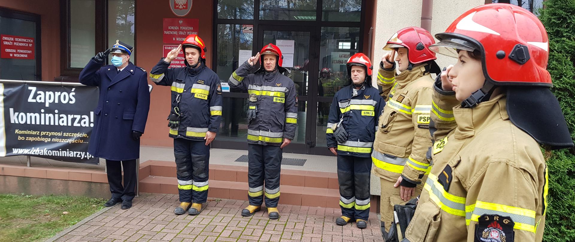 Komendant Powiatowy Państwowej Straży Pożarnej w Wołominie bryg. Andrzej Wysocki w umundurowaniu galowym oraz strażacy z Jednostki Ratowniczo - Gaśniczej w Wołominie ubrani w umundurowanie bojowe, ustawieni w szeregu na tle wejścia głównego do Komendy Powiatowej podczas oddawania honoru w trakcie podniesienia Flagi Państwowej z okazji Narodowego Święta Niepodległości. W tle baner z kampanią promocyjną "Zaproś Kominiarza".