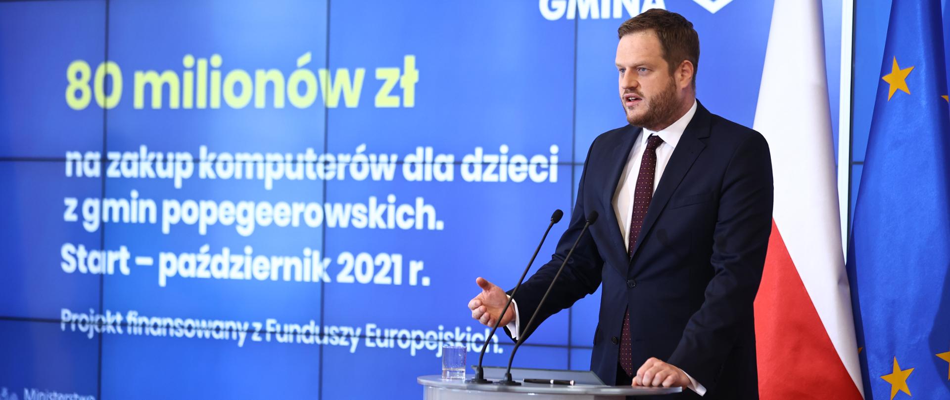 Minister Janusz Cieszyński w trakcie wystąpienia na konferencji prasowej. Stoi przy mównicy z mikrofonami, z lewej strony widoczny ekran prezentacji z tekstem: 80 milionów na zakup komputerów dla dzieci z gmin popegeerowskich. Start - październik 2021. Projekt finansowany z Funduszy Europejskich. Za ministrem flagi Polski i Unii Europejskiej. 