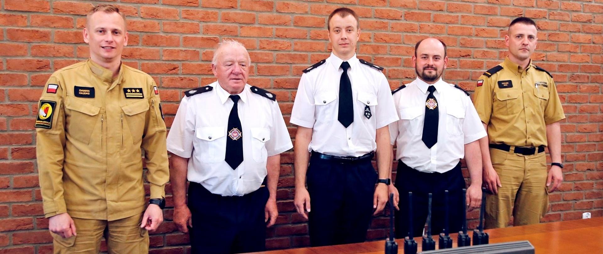 Stoją obok siebie od lewej Zastępca Komendanta Centralnej Szkoły PSP trzech druhów OSP oraz funkcjonariusz wydziału informatyki i łączności CS PSP. Za nimi na ścianie metalowe godło RP przed nimi na blacie przekazywany sprzęt łączności tj. sześć radiotelefonów Motorola GP340 oraz ładowarka sześciostanowiskowa Motorola
