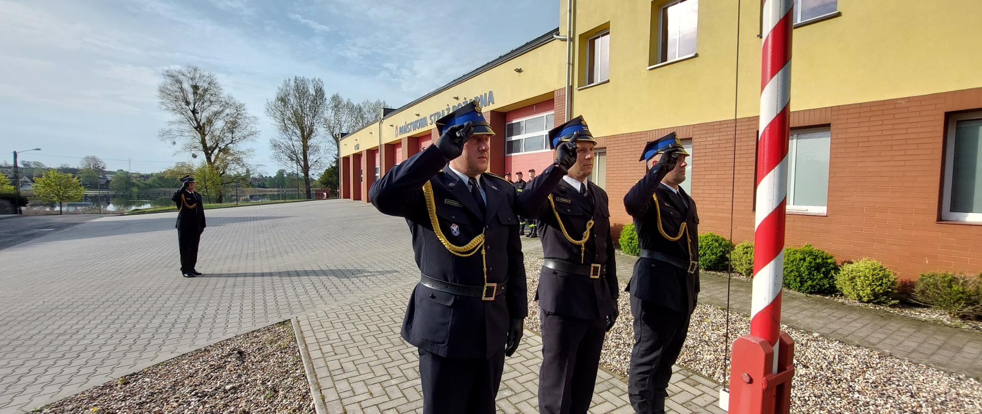 Funkcjonariusze KP PSP Międzychód podczas uroczystego podniesienia Flagi Rzeczpospolitej Polskiej. W tle budynek Komendy Państwowej Straży Pożarnej w Międzychodzie.
