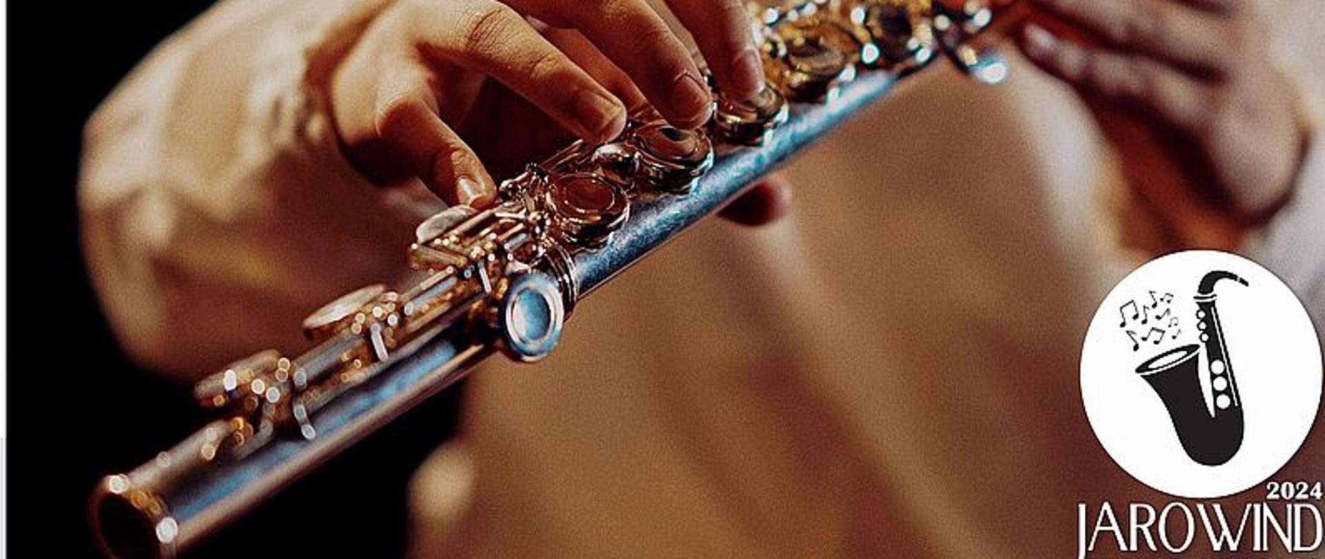 Zdjęcie przedstawia plakat informujący o Regionalnym Przeglądzie Instrumentów Dętych Drewnianych - Jarowind 2024. Na zdjęciu ręce osoby grającej na flecie.
Plakat o szarym tle. 