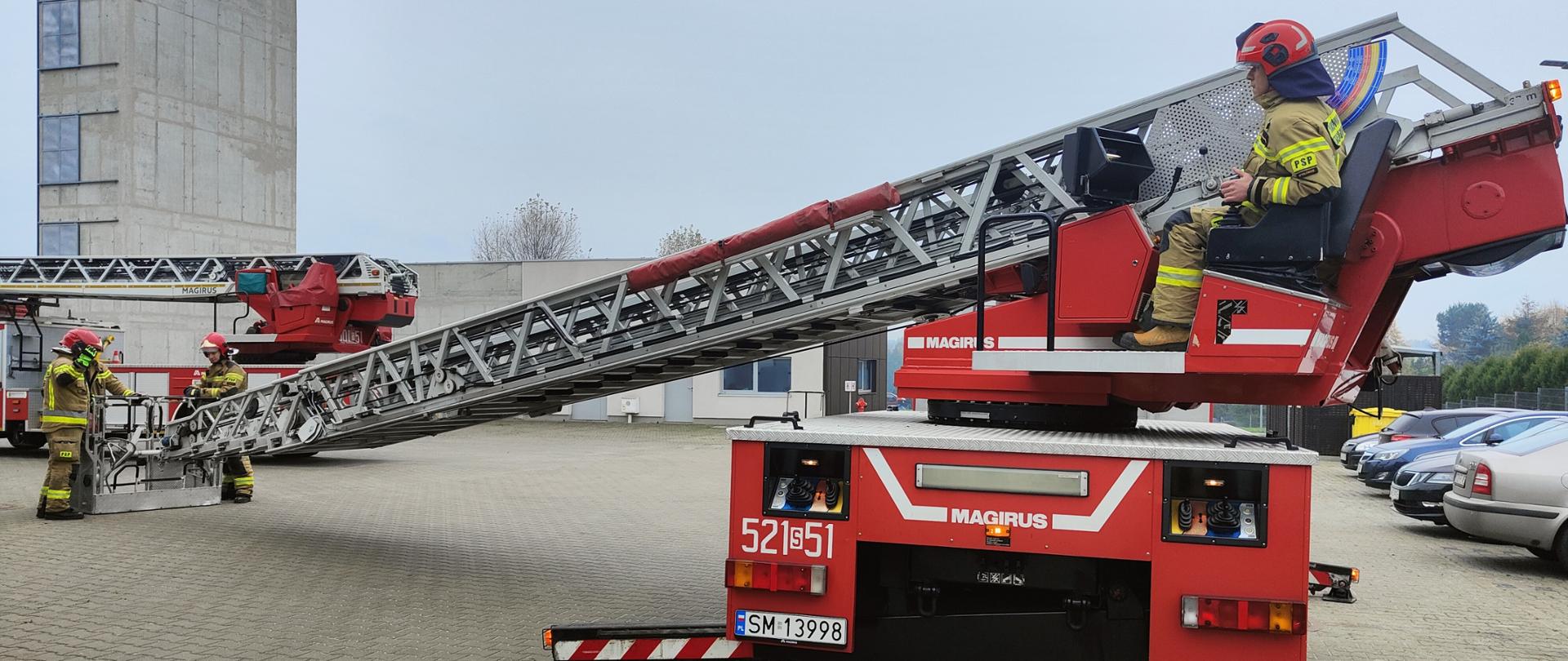 Parking z samochodami. Na pierwszym planie samochód specjalny - drabina w kolorze czerwonym. Na fotelu operatora siedzi strażak w ubraniu specjalnym i hełmie, przesło drabiny wysunięte na bok. Obok kosza drabiny stoi dwóch strażaków w ubraniach specjalnych i hełmach. Za nimi zaparkowany drugi samochód specjalny - drabina.