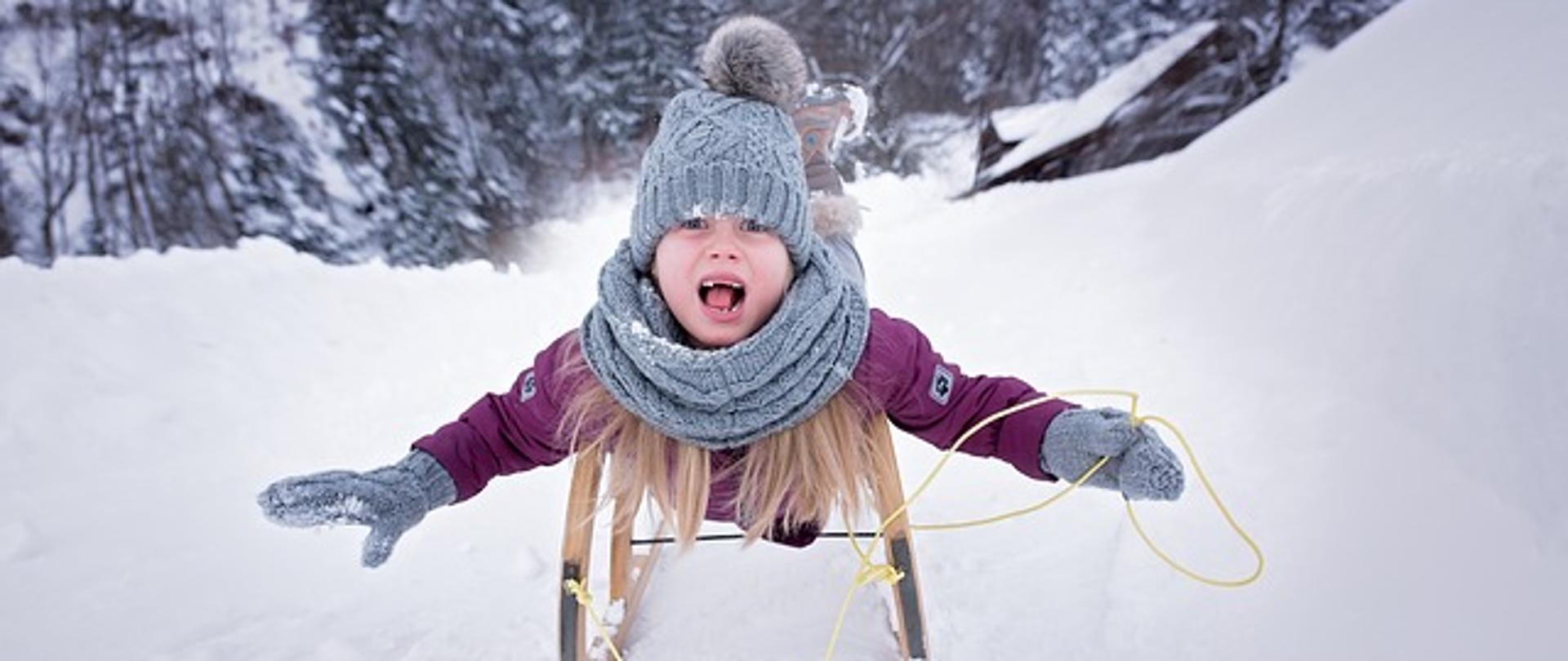 zdjęcie dziewczynki ubranej w kurtkę, czapkę i szalik, zjeżdża na sankach po zaśnieżonej górce. 