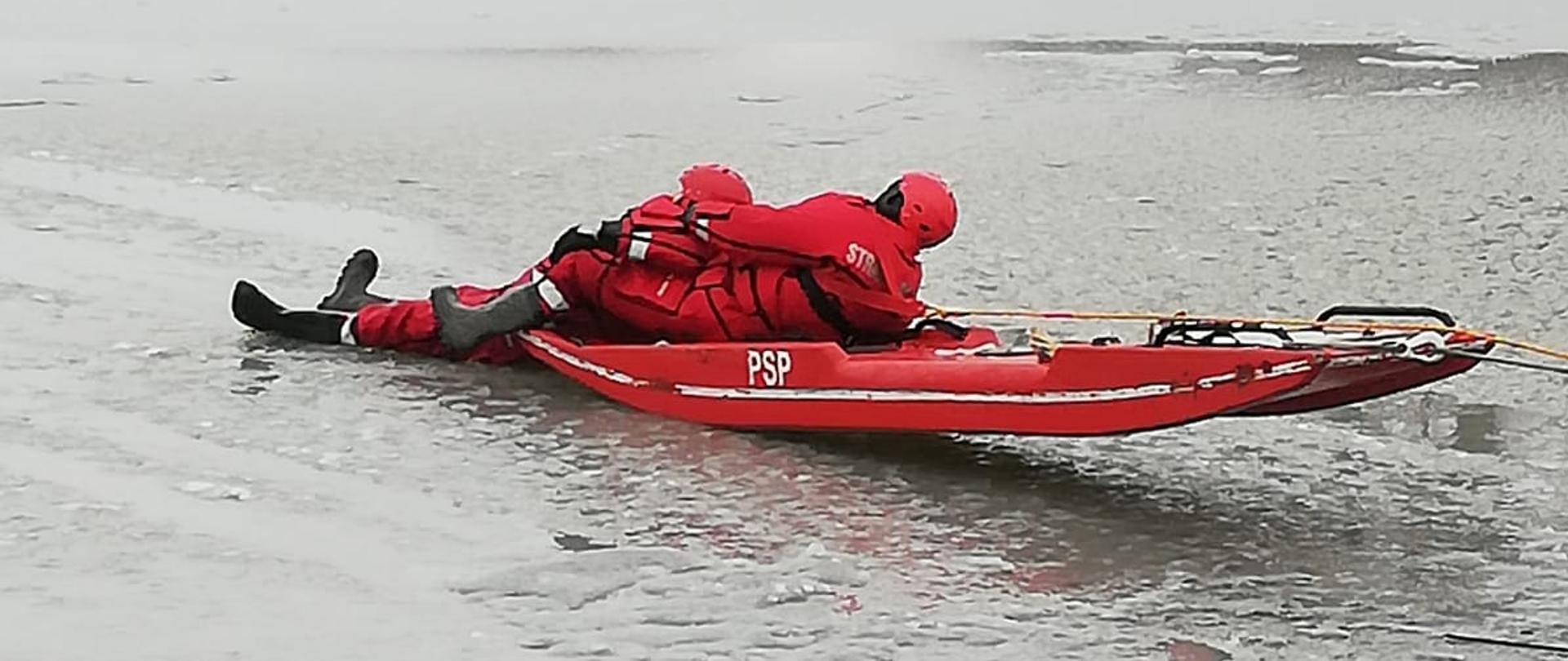 Na zdjęciu strażacy ubrani w czerwone ubrania wypornościowe przy pomocy deskosani ćwiczą ewakuację osoby z zamarzniętego akwenu.