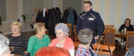 Spotkanie z seniorami. Sala w jednej z rawickich restauracji. Fragment stołu, przy którym po obu stronach siedzą uczestniczki spotkania - seniorki z osiedla Sienkiewicza w Rawiczu . Za stołem stoi policjant wygłaszający pogadankę o zagrożeniach.