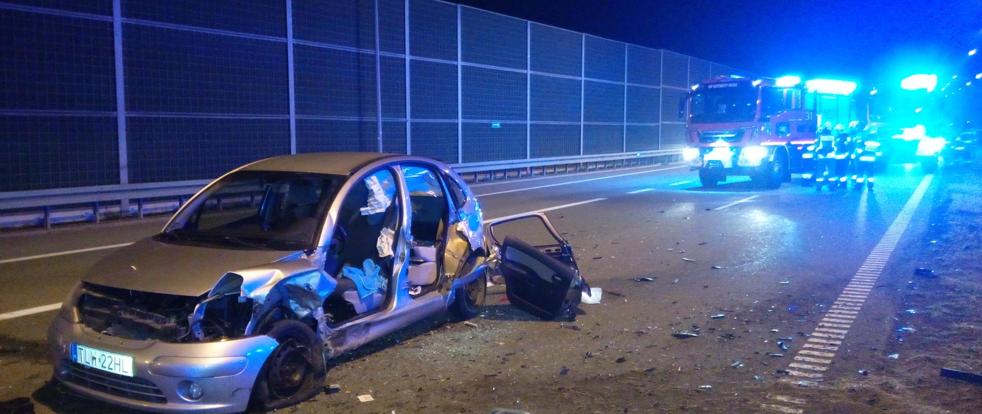Zdjęcie przedstawia miejsce wypadku. Po lewej stronie rozbity Citroen - brak drzwi, do okoła porozrzucane części samochodu. W tle samochody pożarnicze.