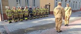 Ćwiczenia w szpitalu Dziedziniec szpitala. Na tle budynku, w dwuszeregu ustawieni sa strażacy uczestniczący w ćwiczeniach Przed nimi zastępca dowódcy JRG składa meldunek komendantowi powiatowemu PSP o zakończeniu ćwiczeń. budynki. 
