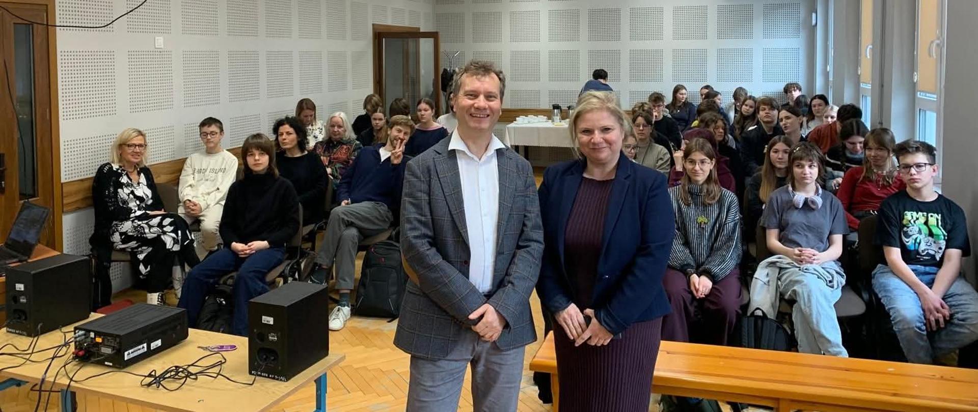 w tle zdjęcia widać osoby siedzące na krzesłach na pierwszym planie dyrektorka szkoły we fioletowej sukience i granatowej marynarce obok niej wykładowca w szarym garniturze i białej koszuli