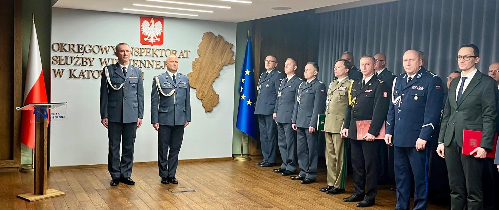 Na zdjęciu widać uroczystą scenę w sali reprezentacyjnej. Po lewej stronie stoi mównica z emblematem Służby Więziennej. Obok niej ustawiona jest pionowo polska flaga – biało-czerwona. Nad nią, na ścianie, znajduje się godło Polski: biały orzeł na czerwonym tle. Na środku sali stoją dwaj funkcjonariusze w szarych, galowych mundurach Służby Więziennej. Są zwróceni przodem, stoją prosto z rękami wzdłuż ciała. Za nimi na ścianie widoczny jest duży napis: „Okręgowy Inspektorat Służby Więziennej w Katowicach”. Obok napisu znajduje się duża, drewniana dekoracja w kształcie mapy regionu. Po prawej stronie stoi rząd kilkunastu osób. Większość z nich to mężczyźni w mundurach różnych służb – głównie szarych i granatowych – z odznaczeniami i oznaczeniami stopni. Jeden z nich ma mundur w kolorze oliwkowym, przypominający mundur wojskowy. Wśród nich stoi także śląski komendant wojewódzki Państwowej Straży Pożarnej starszy brygadier Damian Legierski. Kilka osób trzyma w rękach czerwone teczki z godłem Polski. Za grupą ustawiona jest flaga Unii Europejskiej – niebieska z żółtymi gwiazdami. 