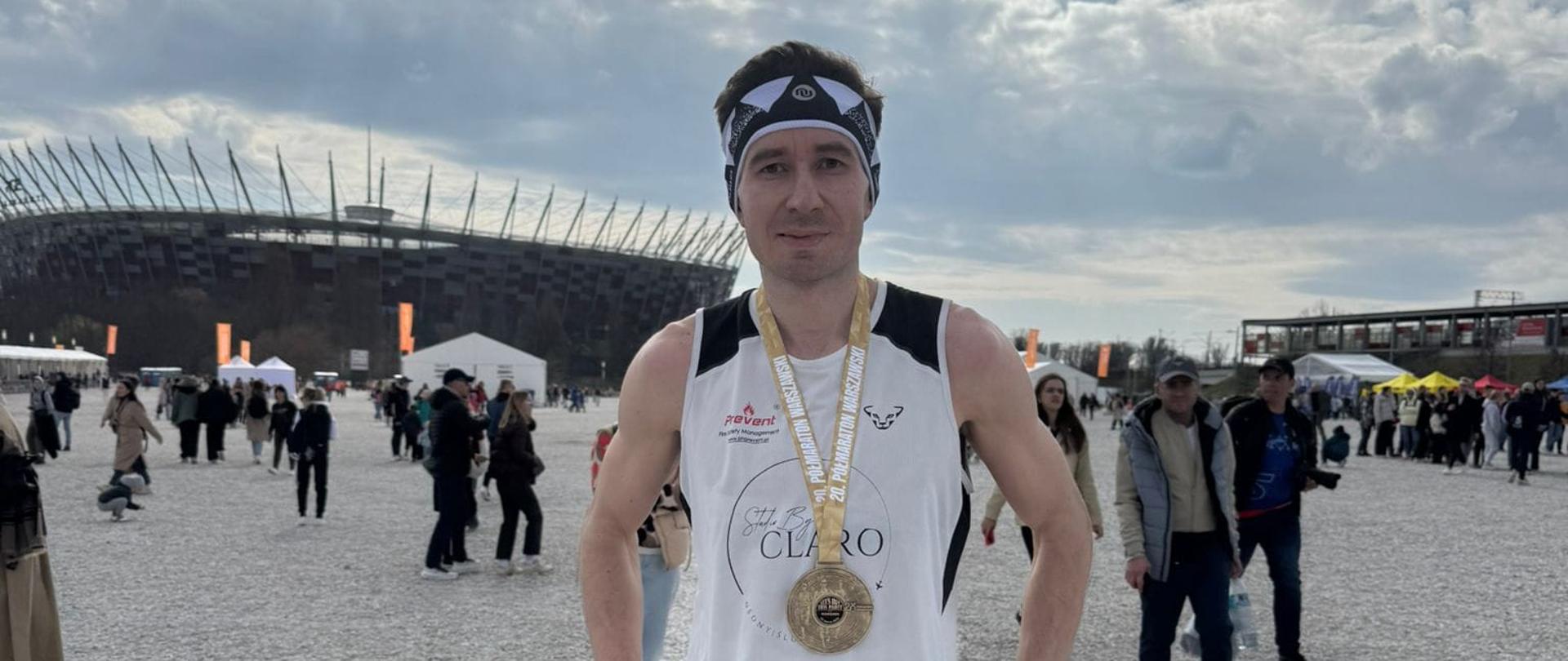 Na planie głównym funkcjonariusz w ubraniu sportowym w tle dostrzegamy zabudowania stadionu w Warszawie. Zdjęcie wykonane po ukończonym starcie na klatce piersiowej medal.