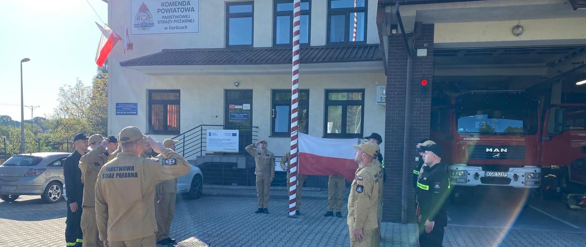 Na zdjęciu widoczne zmiany służbowe w dwuszeregu w pozycji na baczność w ubraniach koszarowych koloru pisakowego i czarnego w czapkach podczas uroczystej zmiany służby w trakcie podnoszenia flagi w zawiązku z uroczystością Dnia Flagi Rzeczypospolitej Polskiej.