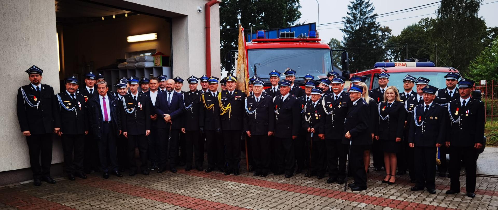 strażacy psp, druhowie osp inni uczestnicy uroczystości na zbiorowym zdjęciu, za zgromadzonymi budynek remizy oraz samochody pożarnicze