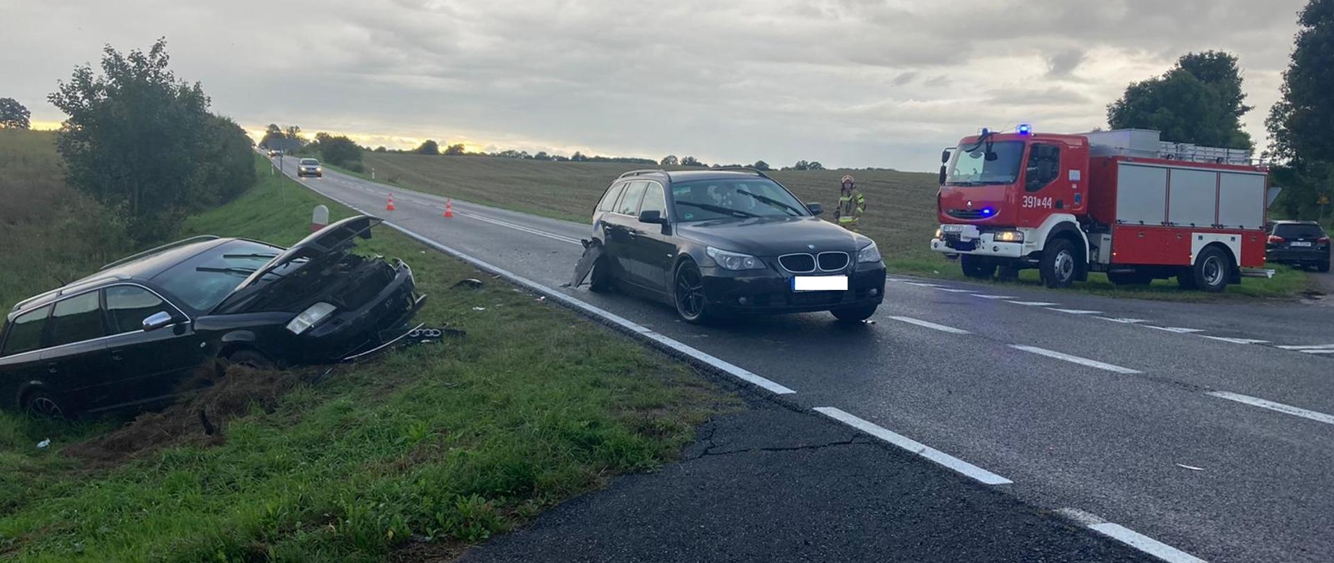Widok stojącego na jezdni uszkodzonego samochodu osobowego, w rowie po lewej stronie kolejny samochód osobowy z uszkodzonym przodem. Po prawej stronie samochód strażacki i stojący obok strażak