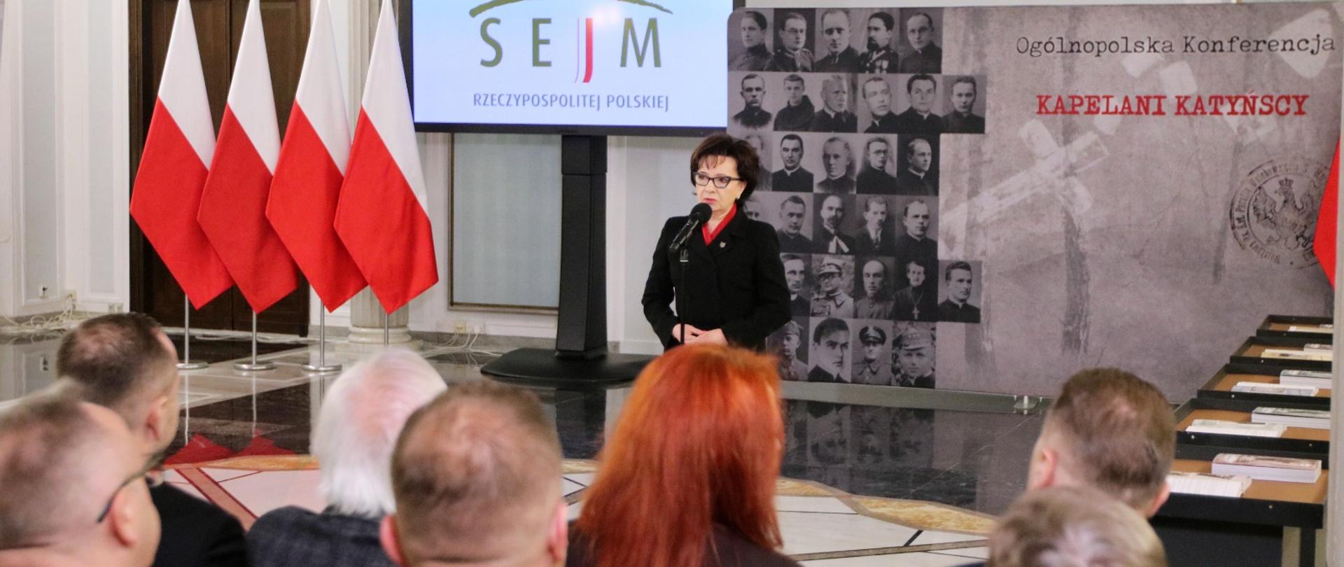 Z przodu sali stoi duży monitor z napisem Sejm, obok niego kobieta w czarnej marynarce mówi do mikrofonu na stojaku, za nią ścianka z napisem Kapelani katyńscy, przed nią na ustawionych w rzędy krzesłach siedzą ludzie.