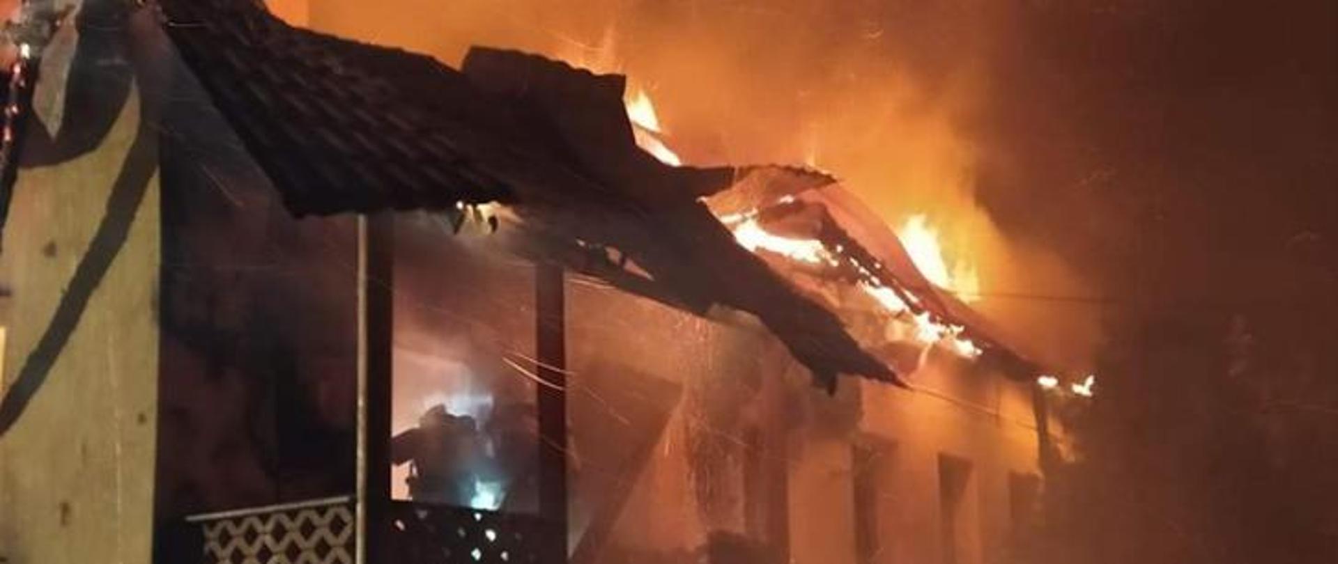 Zdjęcie nocne przedstawiające pożar parterowego budynku mieszkalnego jednorodzinnego. Płomienie wydobywają się spod pokrycia dachu. W wejściu do płonącego budynku widoczny jest strażak w ubraniu specjalnym, wyposażony w aparat powietrzny.