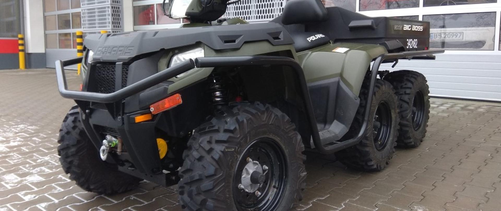 Na pierwszym planie pojazd typu Quad w kolorze ciemno zielonym do przewozu sprzętu pożarniczego w trudno dostępne miejsca. W tle garaże Jednostki Ratowniczo-Gaśniczej nr 2.