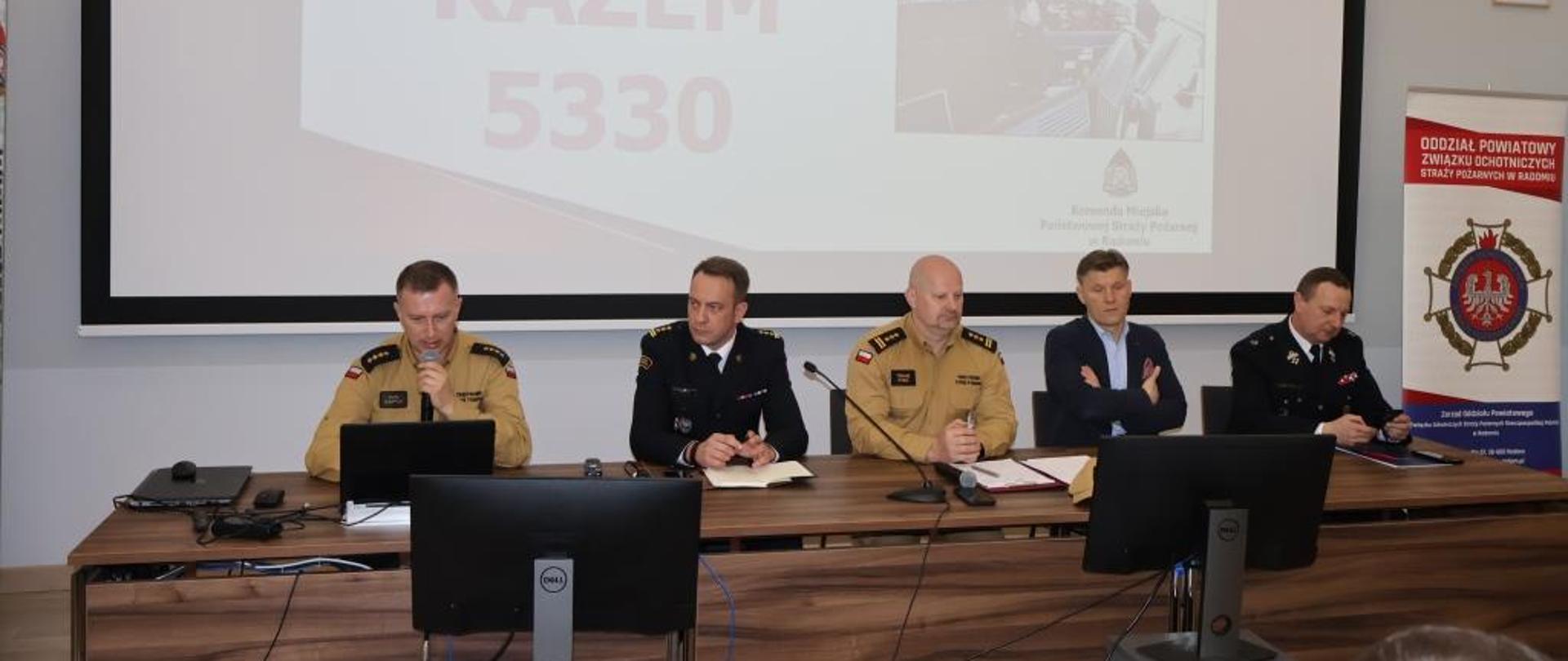 Zdjęcie wykonane w Leśnym Ośrodku Edukacyjnym w Jedni – Letnisku. W obiekcie tym odbyła się Narada Komendanta Miejskiego PSP w Radomiu oraz Prezesa ZOSP RP w Radomiu. Na zdjęciu widać w kolejności od lewej: naczelnika wydziału operacyjnego KM PSP w Radomiu starszego kapitana Piotra Chłopickiego. Funkcjonariusz ubrany w ubranie służbowe w kolorze żółtym piaskowym. W ręku trzyma mikrofon przez który referuje prezentację. Obok niego siedzą dalej: Komendant Miejski PSP w Radomiu starszy brygadier Bartosz Musiał, zastępca Mazowieckiego Komendanta Wojewódzkiego PSP starszy brygadier Tomasz Cybul, starosta powiatu radomskiego Waldemar Trelka oraz Prezes Zarządu Oddziału Powiatowego ZOSP RP w Radomiu druh Paweł Tuzinek. 