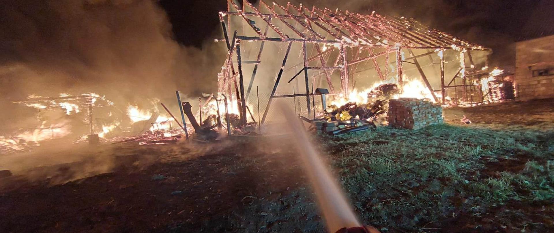 Zdjęcie przedstawia strażaków gaszących pożar stodoły.
W tle inne budynki.
