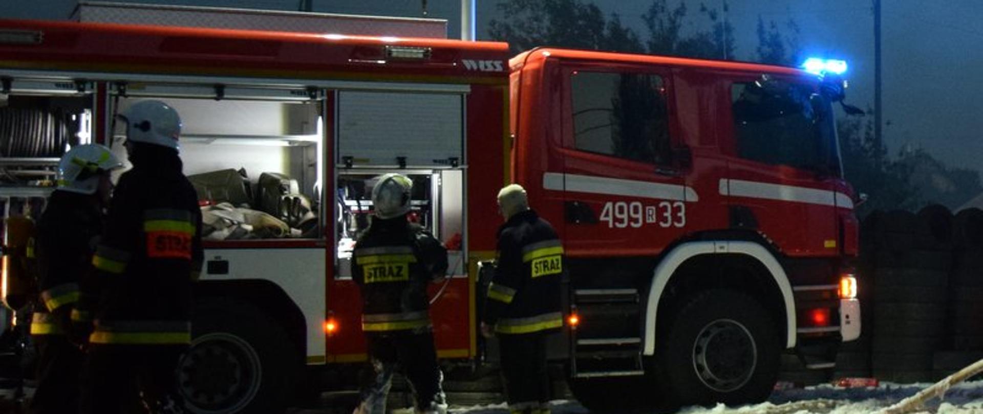 Zdjęcie zrobione w nocy. Na zdjęciu widać samochód gaśniczy, który ma włączone sygnały błyskowe. Przed samochodem stoją strażacy w ubraniach specjalnych (nomexach), przygotowujący się do gaszenia pożaru.