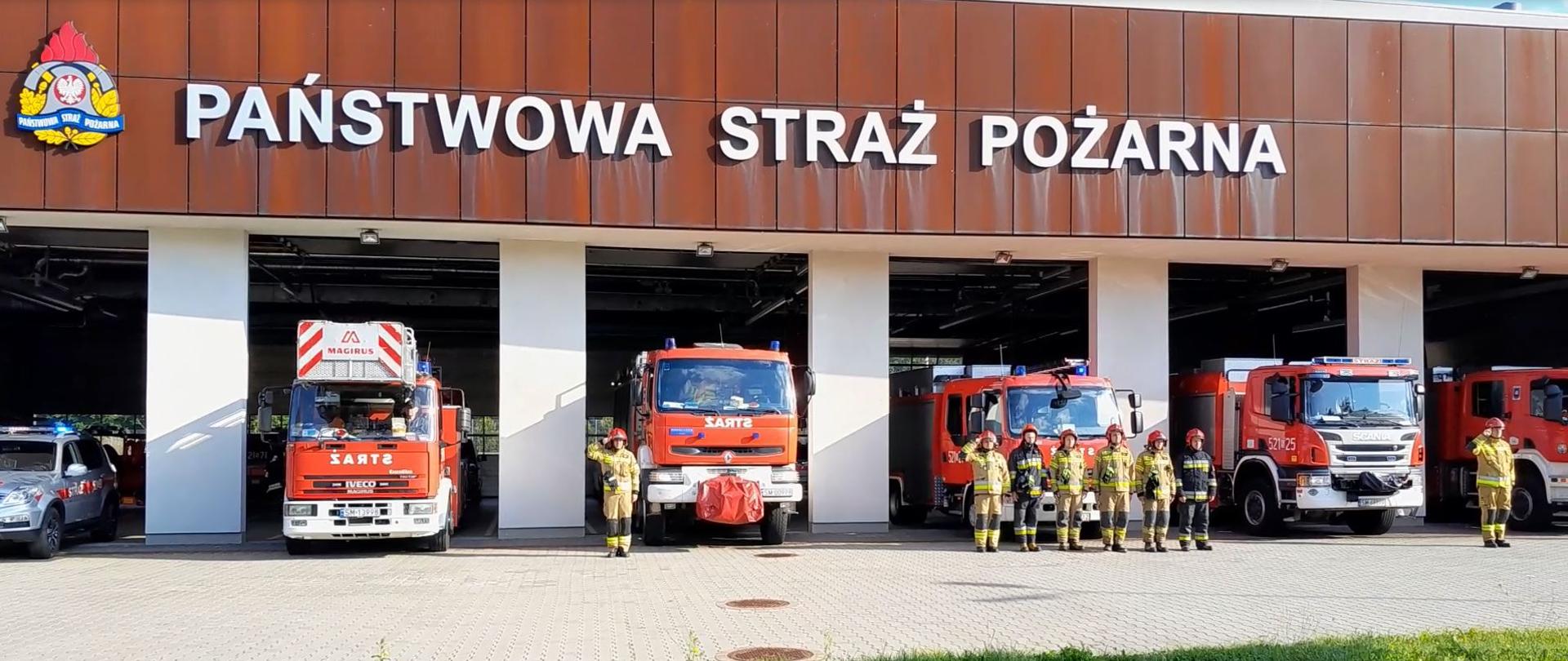 Strażacy składający hołd funkcjonariuszowi Komendy Głównej PSP, który zginął w wypadku podczas służby na tle samochodów bojowych stojących w bramach wyjazdowych komendy miejskiej.