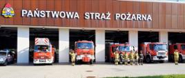 Strażacy składający hołd funkcjonariuszowi Komendy Głównej PSP, który zginął w wypadku podczas służby na tle samochodów bojowych stojących w bramach wyjazdowych komendy miejskiej.