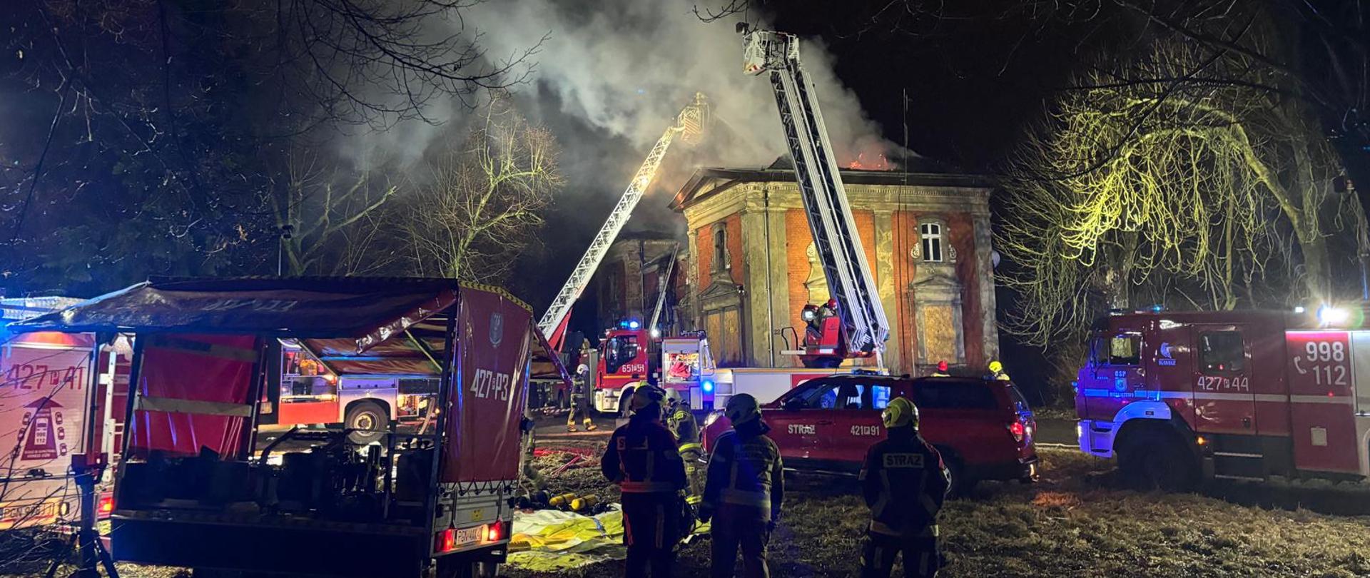 Na zdjęciu widać strażaków gaszących pożar dużego, zabytkowego budynku nocą. Z dachu unosi się gęsty dym, a dwie drabiny strażackie są skierowane w stronę płomieni. Teren oświetlają światła wozów strażackich, wokół stoją ratownicy i sprzęt gaśniczy.