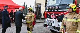na pierwszym planie dwóch strażaków ubranych w piaskowych ubraniach stoi przy samochodzie pożarniczym na którym założona jest wstęga, dwie osoby jedna w mundurze strażackim oraz ubraniu cywilnym dokonują przekazania dokumentów oraz kluczyków do w/w samochodu 