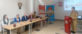 Zdjęcie pokazuje st. bryg. Wojciecha Szczepanika Komendanta Powiatowego Państwowej Straży Pożarnej w Gołdapi, który rozpoczyna przedstawianie w formie prezentacji multimedialnej podsumowania zadań realizowanych przez Komendę Powiatową Państwowej Straży Pożarnej w Gołdapi w 2024 roku oraz planów na rok 2025. Z lewej strony obrazu widać również obecnych na sali zaproszonych gości